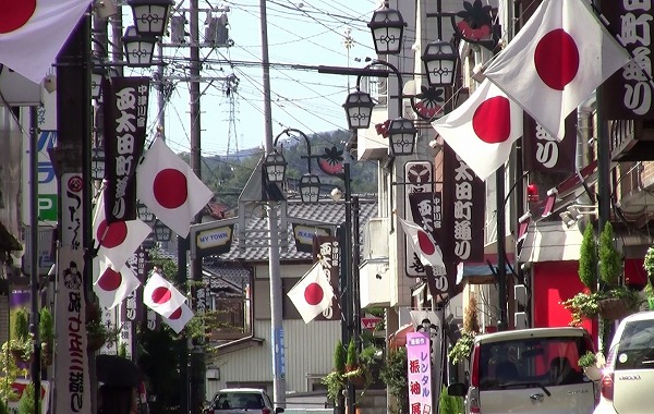 Na imagem vemos uma rua de uma cidade do Japão cheia de prédios dos dois lados, com vários carros e também postes na rua. Em praticamente todos os prédios podemos ver uma bandeira do Japão hasteada, que é branca com um circulo vermelho, em comemoração.
