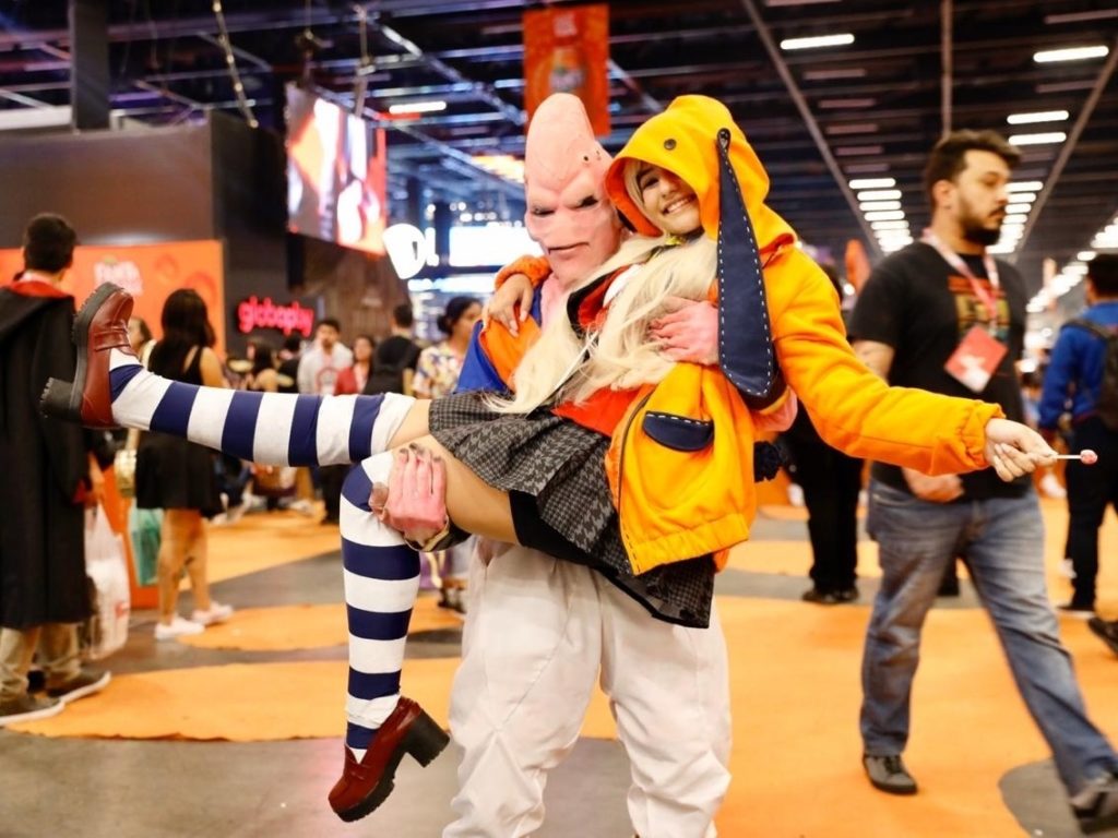 Um cosplay do personagem Majin Boo de Dragon Ball Z, segurando uma mulher cosplayer no colo, ambos posando para uma foto em um evento geek.