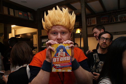 Homem fantasiado de Goku, personagem da franquia Dragon Ball, segurando um livro com uma capa escrito The Otaku Encyclopedia.
